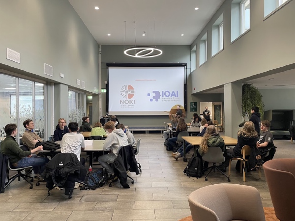 NOKI åpningsseremoni. NOKI og IOAI-logo på storskjerm, deltakere sitter rundt bord.